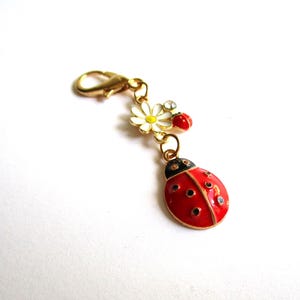 Op de afbeelding: Een goudkleurige metalen bedel met een grote rood-zwarte geëmailleerde lieveheersbeestjeshanger, een kleine witte madelief met een geel hartje en een minuscuul rood lieveheersbeestje. De bedel is voorzien van een karabijnsluiting voor bevestiging.
