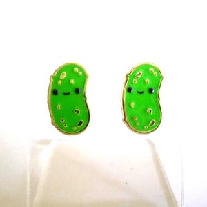 May include: A pair of green pickle-shaped earrings with gold-colored trim. Each earring features a smiling face with black eyes and a small mouth. The earrings are on a white background.