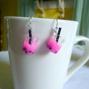 Op de afbeelding: Een paar oorbellen met miniatuur roze bubble tea bekers met zwarte rietjes. De oorbellen zijn zilver en hebben een hangend ontwerp.