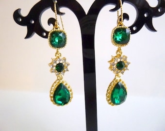May include: A pair of gold tone dangle earrings with emerald green and clear rhinestones. The earrings have a teardrop shape with a smaller square stone at the top.