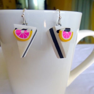 May include: A pair of silver dangle earrings with a white, black, and pink cake slice charm. The cake slice has a pink and yellow citrus fruit on top.