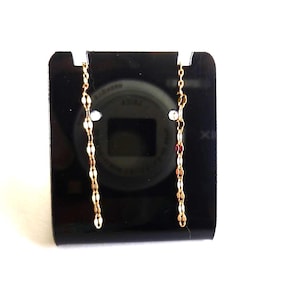 May include: A pair of gold-toned chain earrings with a delicate, elongated design. The earrings are displayed on a black stand against a white background. The chain links alternate between gold and a lighter, possibly silver, color.