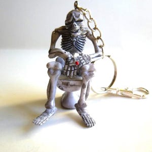 May include: A novelty keychain featuring a small, detailed skeleton figure seated on a miniature toilet. The skeleton is gray and white with a red gem on its hand. A silver chain and keyring are attached.