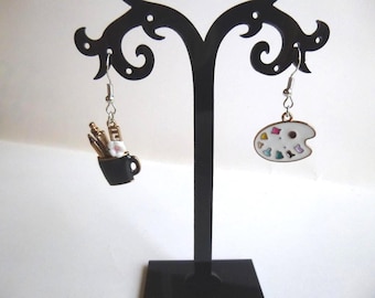May include: A pair of artisan earrings. One earring features a black mug with paintbrushes and pencils. The other earring is a white artist's palette with colorful paint splotches. Both earrings hang from silver hooks.