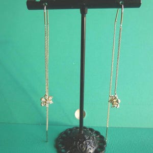 May include: A pair of gold-coloured chain earrings with a floral design at the bottom. The earrings are displayed on a black earring stand against a green background. The floral design is encrusted with small, clear stones.