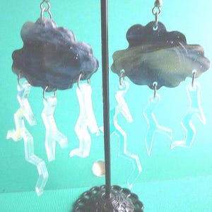 May include: A pair of unique earrings featuring dark gray cloud shapes with clear, lightning bolt charms dangling below. The earrings are suspended from silver-colored hooks, creating a whimsical accessory.