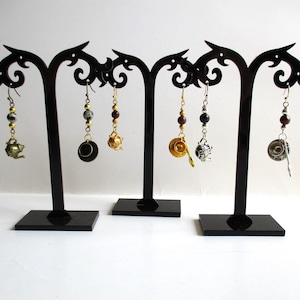 May include: Six dangling earrings featuring miniature tea-themed charms, including teapots, cups, and saucers in gold and silver tones. Each earring is accented with small round beads and hangs from a hook, displayed on three black stands.