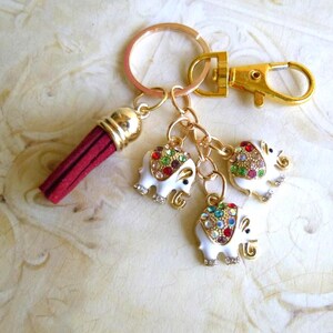 May include: A gold-toned keychain with a red tassel and three white elephant charms. Each elephant charm is adorned with colorful rhinestones. The keychain also features a gold-colored clasp.