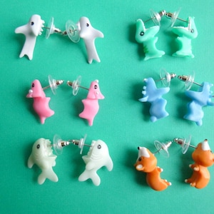 Puede incluir: Un conjunto de pendientes de fantasía con diseños de criaturas marinas. Los pendientes, en pares, presentan diseños de tiburones, delfines y otros animales marinos en blanco, rosa, verde, azul y naranja. Cada pendiente tiene un poste y un cierre transparente.