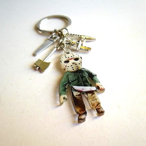 May include: A silver keychain with multiple charms. The charms include a small figure of a character wearing a hockey mask and holding a bloody knife, along with miniature saw, axe, and other tool charms. The figure is wearing a green jacket and brown pants.
