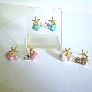 Puede incluir: Pendientes con diseño de concha y estrella de mar. Los pendientes están disponibles en azul, rosa y blanco, con detalles dorados. Cada pendiente presenta una perla o una gema transparente. Los pendientes se exhiben en un soporte transparente.