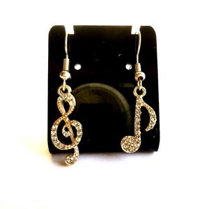 May include: A pair of gold-colored earrings, one shaped like a treble clef and the other like a musical note. Both earrings are encrusted with small, clear crystals. The earrings hang from silver-colored hooks and are displayed on a black background.