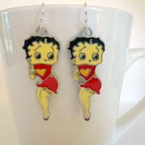 Op de afbeelding: Een paar zilveren oorbellen met een cartoonfiguur met zwart haar, een geel gezicht, een rode jurk en een hartvormig ontwerp. De oorbellen hangen aan een witte mok.