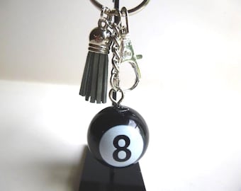 May include: A black and white 8-ball keychain with a silver-tone chain, clasp, and tassel. The keychain features a small, dark gray tassel and a round, glossy black 8-ball with a white number 8.