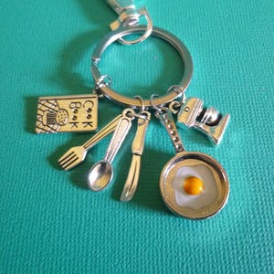 May include: Silver-toned keychain with cooking-themed charms. Charms include a cookbook, fork, spoon, knife, frying pan with a fried egg, and a mixer. The cookbook charm has the word "Cook Book" on it.