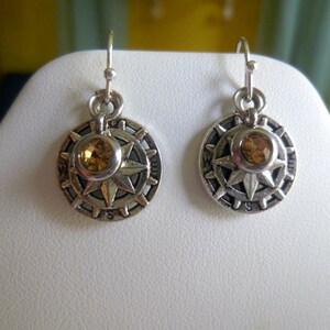 May include: A pair of silver earrings with a compass design and a small amber-colored gemstone in the center of each earring.