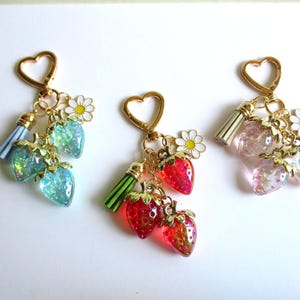 May include: Three heart-shaped gold keychains, each featuring two translucent strawberry charms with glitter, a small white daisy charm with a yellow center, and a single tassel. The keychains come in light blue, bright red, and light pink color variations.