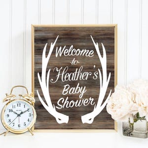 May include: A rustic wooden sign with a gold frame featuring a white antler design and the text "Welcome to Heather's Baby Shower".