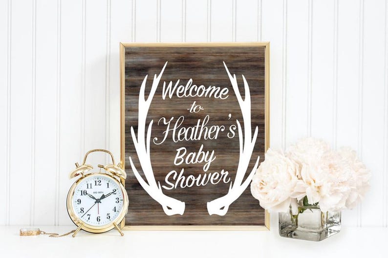 May include: A rustic wooden sign with a gold frame featuring a white antler design and the text "Welcome to Heather's Baby Shower".