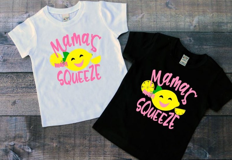 May include: Two white and black t-shirts with a lemon graphic and the text "Mama's Squeeze" in pink.