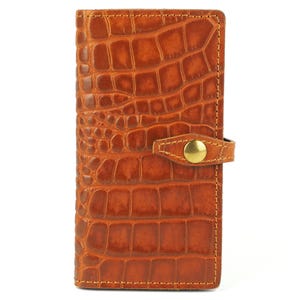 Puede incluir: Cartera de cuero marrón con textura de piel de cocodrilo. Tiene un cierre de botón a presión con un botón dorado. Los bordes están cosidos con hilo marrón claro. La cartera es rectangular y parece cerrada.