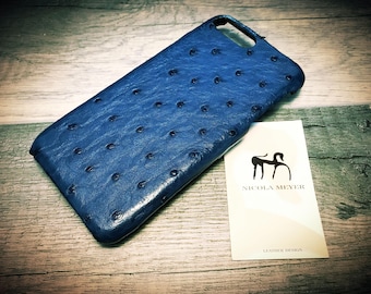 Pode incluir: Uma capa de telefone de couro azul com uma superfície texturizada e pequenas manchas escuras. Um cartão de visita com um logotipo e o texto "NICOLA MEYER LEATHER DESIGN" está ao lado da capa. A capa está em uma superfície de madeira.