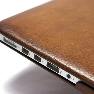 May include: A brown leather laptop case with a silver trim and ports on the side.