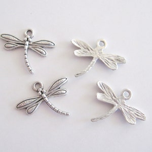 May include: Four silver-colored dragonfly charms. Each charm has detailed wings, a segmented body, and a small loop at the top for attaching to jewelry or other crafts. The charms are on a white background.