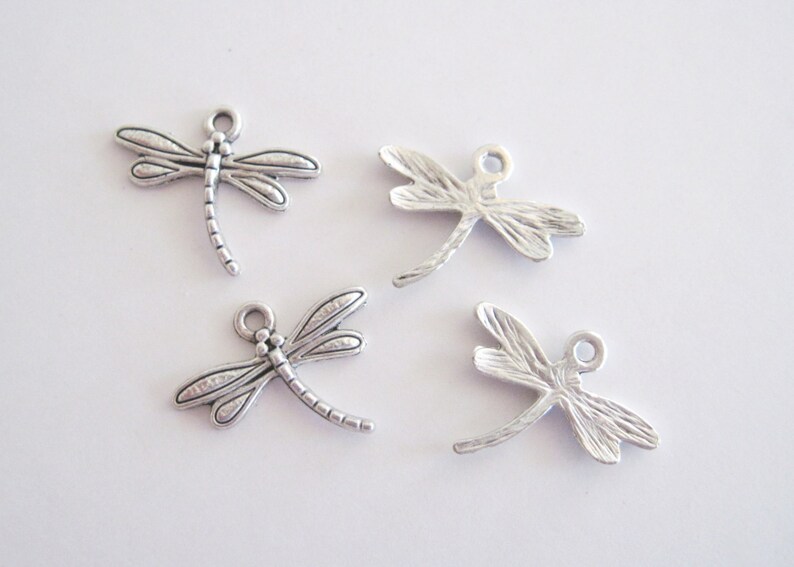May include: Four silver-colored dragonfly charms. Each charm has detailed wings, a segmented body, and a small loop at the top for attaching to jewelry or other crafts. The charms are on a white background.