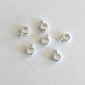 May include: A collection of small, silver-colored pendant bails. Each bail features a circular loop with a small, attached loop for stringing. The bails are scattered on a white surface.