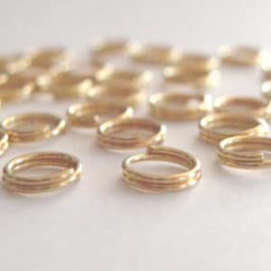 May include: A collection of small, gold-colored metal rings scattered on a white surface. The rings have a spiral design, with multiple coils creating a layered effect. The lighting highlights the metallic sheen of the rings.