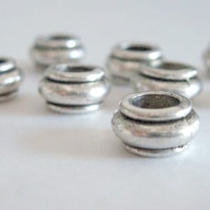 May include: A collection of silver-colored metal beads. Each bead has a rounded, barrel-like shape with a central hole and horizontal ridges. The beads are scattered across a white surface, with some in focus and others slightly blurred.