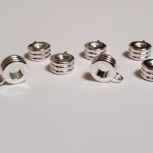 May include: A collection of eight small, shiny silver metal components. Each component has a cylindrical shape with three horizontal grooves and a small loop on one side. The objects are arranged on a white surface.