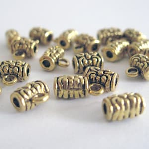 May include: A collection of small, gold-colored metal beads. Each bead is cylindrical with an ornate, textured design and a small loop for stringing. The beads are scattered on a white surface, creating a visually appealing arrangement.
