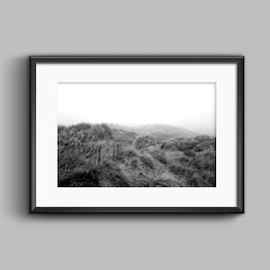 May include: Black and white framed print of a coastal landscape. The image features tall grasses and a fence line on a dune, with a misty sky in the background. The artwork is displayed in a black frame with a white mat.