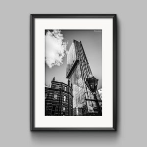 May include: Black and white photograph of a tall modern building in the city. The building is made of glass and has a unique design. A smaller brick building is in the foreground, and a street lamp is visible.