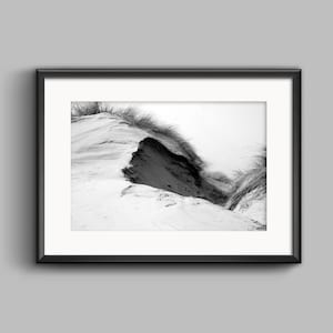 Puede incluir: Una fotografía en blanco y negro de una duna de arena con hierba creciendo en la cima. La duna está parcialmente erosionada, revelando una capa de arena más oscura debajo.