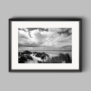Puede incluir: Fotografía en blanco y negro de una escena de playa con dunas de arena y un cielo nublado. La foto está enmarcada en un marco negro.