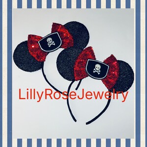 May include: Two black sequined mouse ear headbands with red sequined bows. Each bow features a pirate flag emblem with a skull and crossbones. The text "LillyRoseJewelry" is in red script at the bottom.