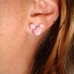 Op de afbeelding: Een close-up van iemands oor met een roze, glinsterende Mickey Mouse oorbel.
