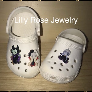 May include: Two white clogs with Disney villain character shoe charms. The charms are of Maleficent, Cruella de Vil, and Ursula. The charms are attached to the front of the clogs. The text "Lilly Rose Jewelry" is visible above the clogs.