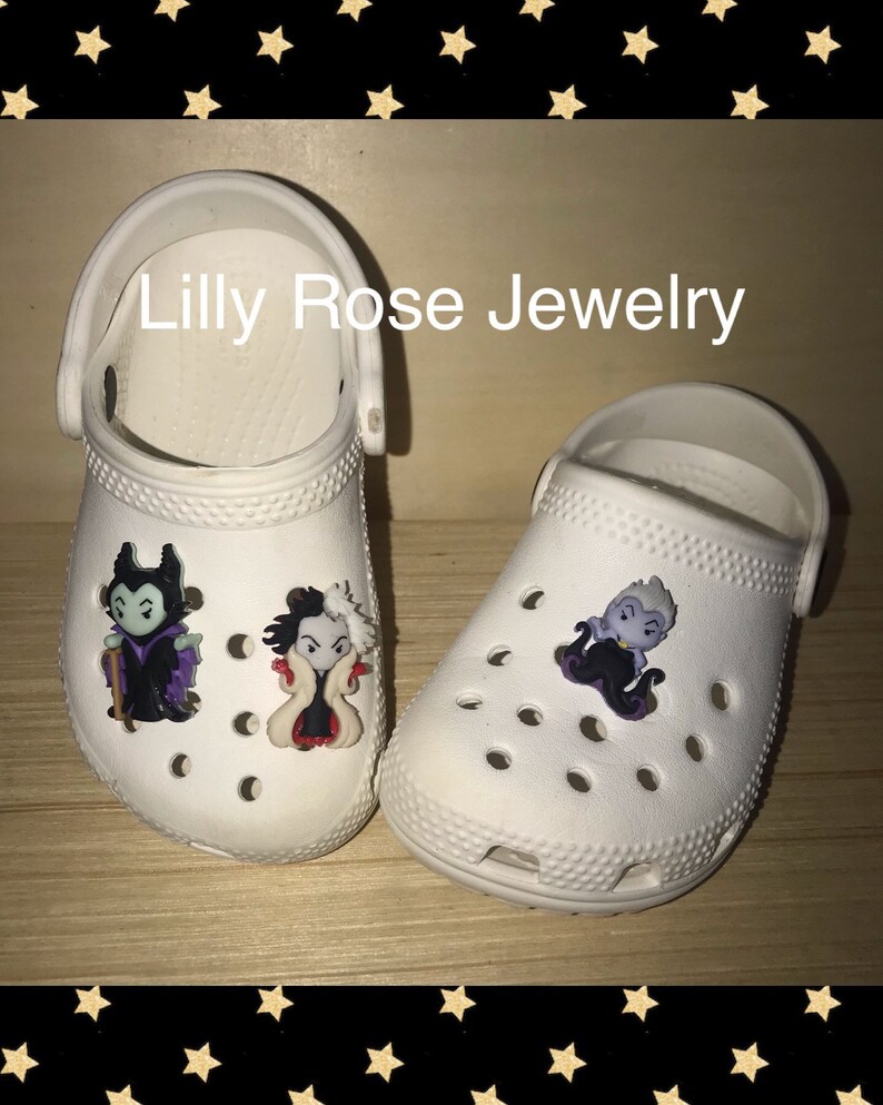 May include: Two white clogs with Disney villain character shoe charms. The charms are of Maleficent, Cruella de Vil, and Ursula. The charms are attached to the front of the clogs. The text "Lilly Rose Jewelry" is visible above the clogs.
