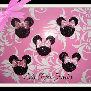 May include: Five black and pink glitter Minnie Mouse head embellishments with pink bows. The embellishments are on a pink background with a white floral pattern. The text "Lilly Rose Jewelry" is at the bottom of the image.