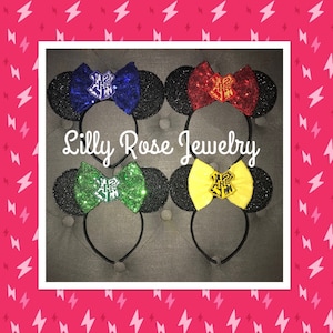 May include: Four pairs of black mouse ears with colorful glitter bows featuring the Hogwarts house crests. The bows are blue, red, green, and yellow. The text "Lilly Rose Jewelry" is printed below the ears.