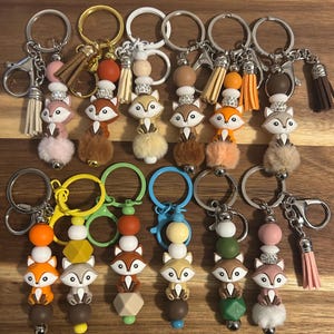 May include: A collection of fox-themed keychains with various colors and designs. Each keychain features a fox charm, decorative beads, and a tassel. The keychains have silver, gold, and colored metal rings and clasps.