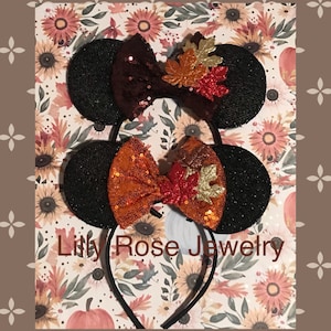 May include: Black mouse ears with a brown and orange sequined bow and a fall leaf embellishment. The headband is black. The text "Lilly Rose Jewelry" is visible in the image.