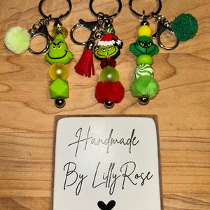 May include: Three handmade keychains featuring Grinch-themed charms, beads, and pom-poms in green, red, and yellow. Each keychain has a silver clasp and ring. A small wooden sign with the text "Handmade By Lilly Rose" and a heart is at the bottom.