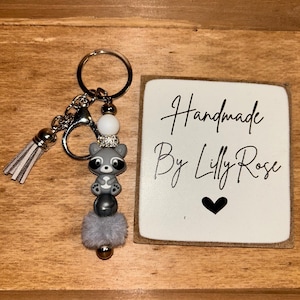 Op de afbeelding: Een handgemaakte sleutelhanger met een grijze wasbeerbedel, zilverkleurige hardware en een pluizige grijze pompon. De sleutelhanger bevat een witte kwast en een klein wit label met de tekst "Handmade By Lilly Rose" en een zwart hart.