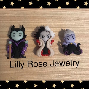 May include: Three small, cartoon villain figurines. The figurines are of Maleficent, Cruella de Vil, and Ursula. The figurines are all in a standing position and are made of a colourful plastic material. The figurines are on a wooden surface with a black and gold star pattern background. The text "Lilly Rose Jewelry" is printed below the figurines.