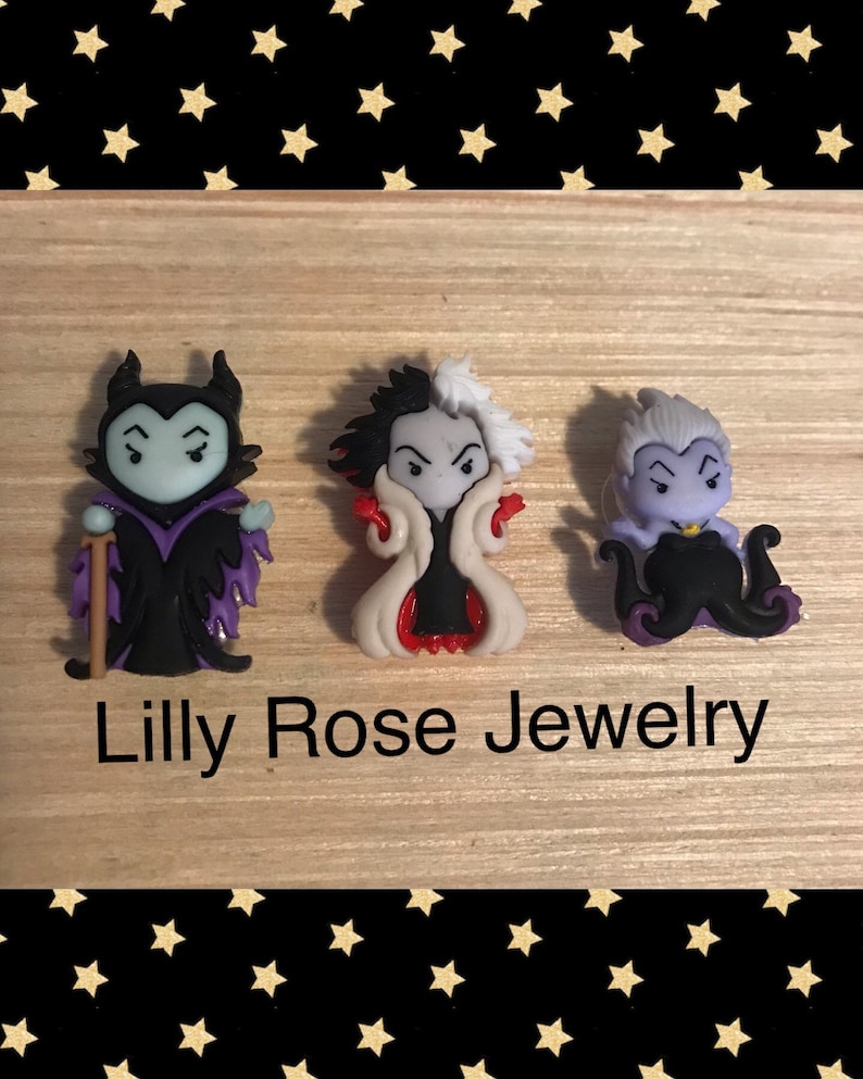May include: Three small, cartoon villain figurines. The figurines are of Maleficent, Cruella de Vil, and Ursula. The figurines are all in a standing position and are made of a colourful plastic material. The figurines are on a wooden surface with a black and gold star pattern background. The text "Lilly Rose Jewelry" is printed below the figurines.